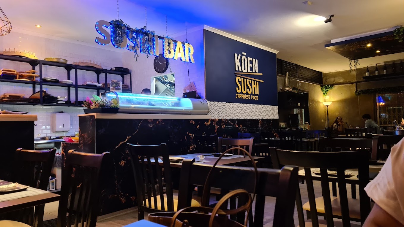 Sala do Koen Sushi à noite, com o letreiro Sushi Bar iluminado e o logótipo na parede azul